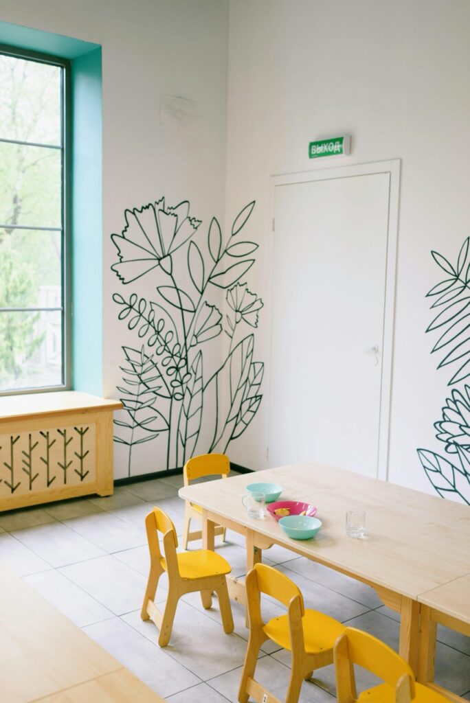 a clean room with white walls with a flower pattern on them. a simple wood table with yellow chairs